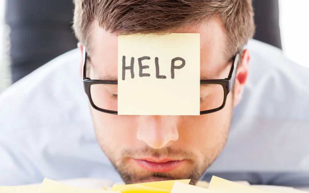 person with their chin on a desk and a post-it note on their forehead reading 'help'
