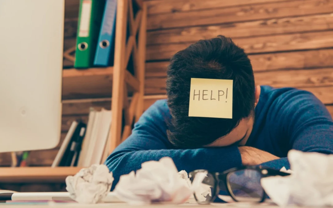 Person at cluttered desk with head down and post-it on head saying "help"