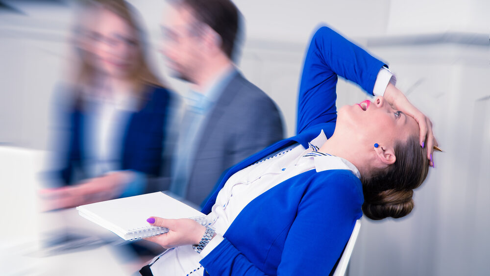 Woman in a meeting leaning way back in her chair with her hand on her head while others in the background talk to each other.