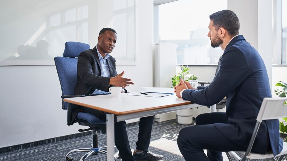 Two business men sitting across a desk from one another having a conversation