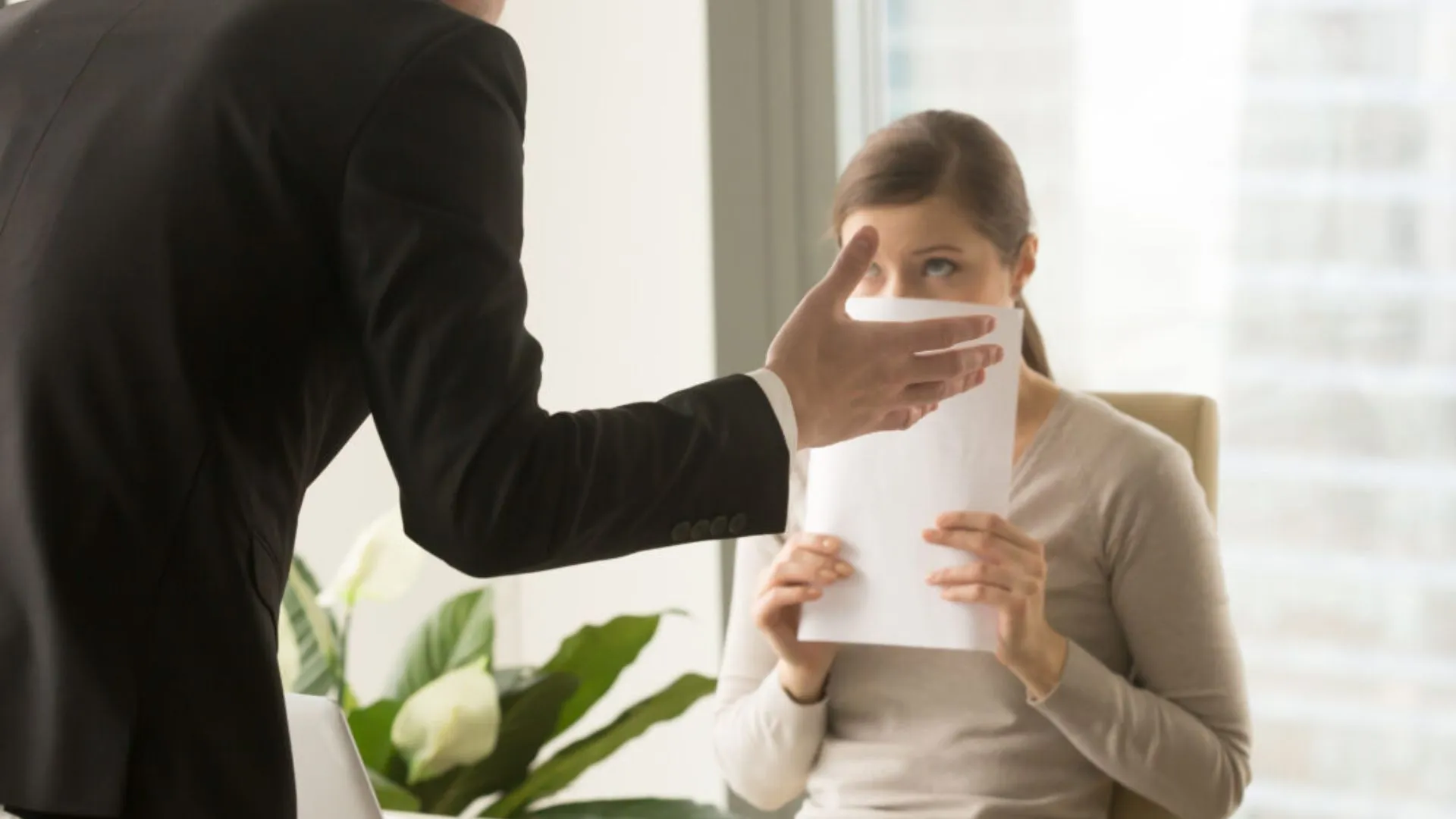 Woman covering her face with a paper while her boss leans over her desk in an intimidating way