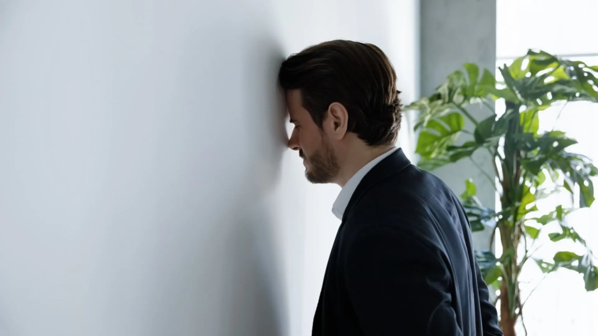 Man leaning his head against a white office wall dealing with too much work stress.