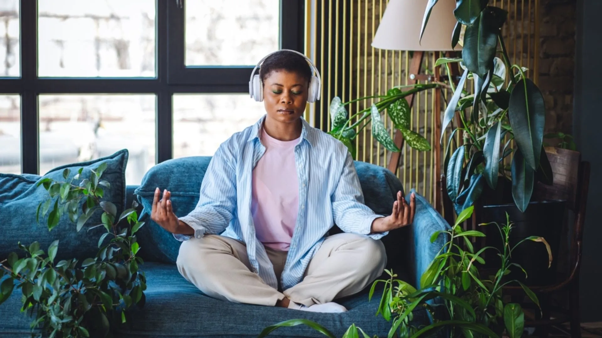 Woman wearing headphones and meditating in a plant-filled room to alleviate stress spillover
