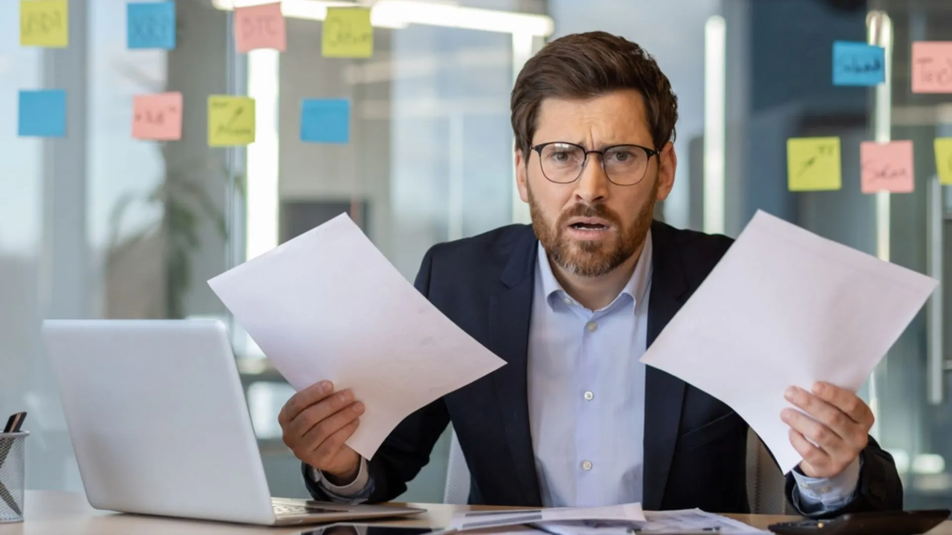 Business man at desk looking at two different pieces of paper with dozens of sticky notes on the window behind him.