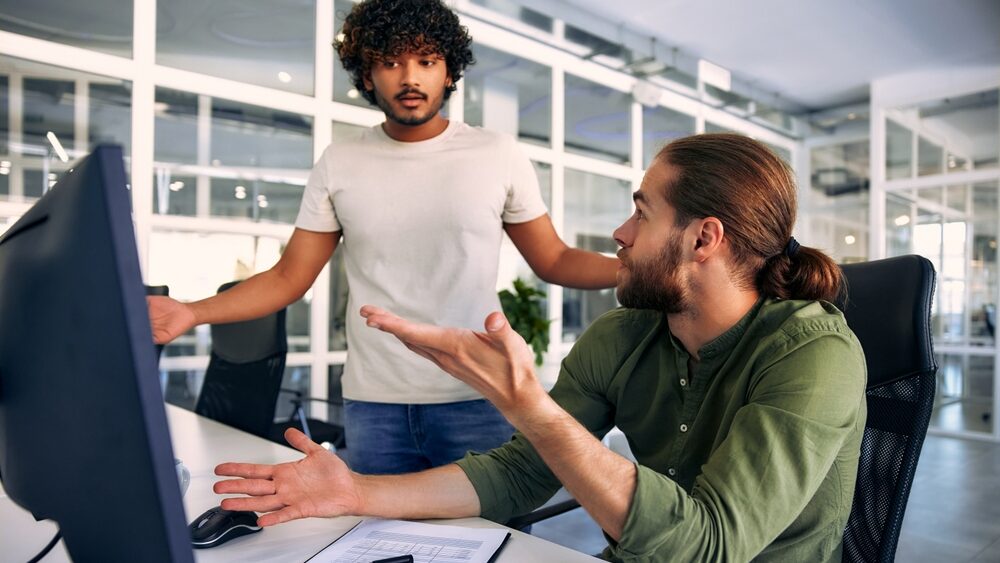 Two colleagues arguing over priorities. One is sitting at a desk, the other is standing over him