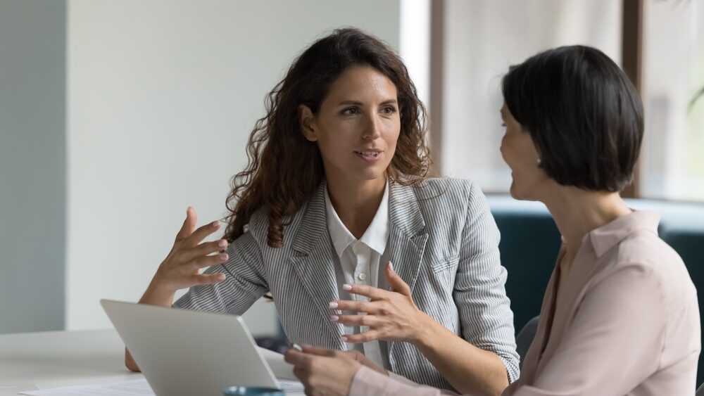 Two women having a conversation at a desk. One is talking and animiating with her hands.