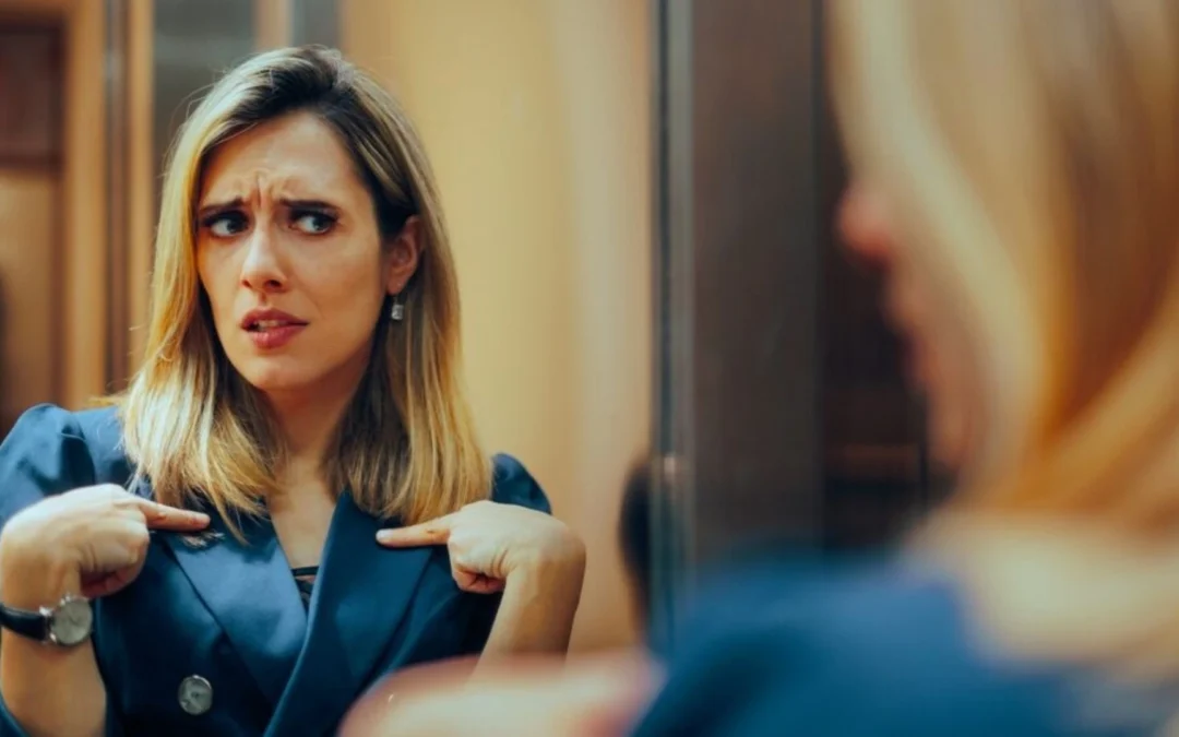 Woman looking in the mirror pointing at herself, questioning if it's about her